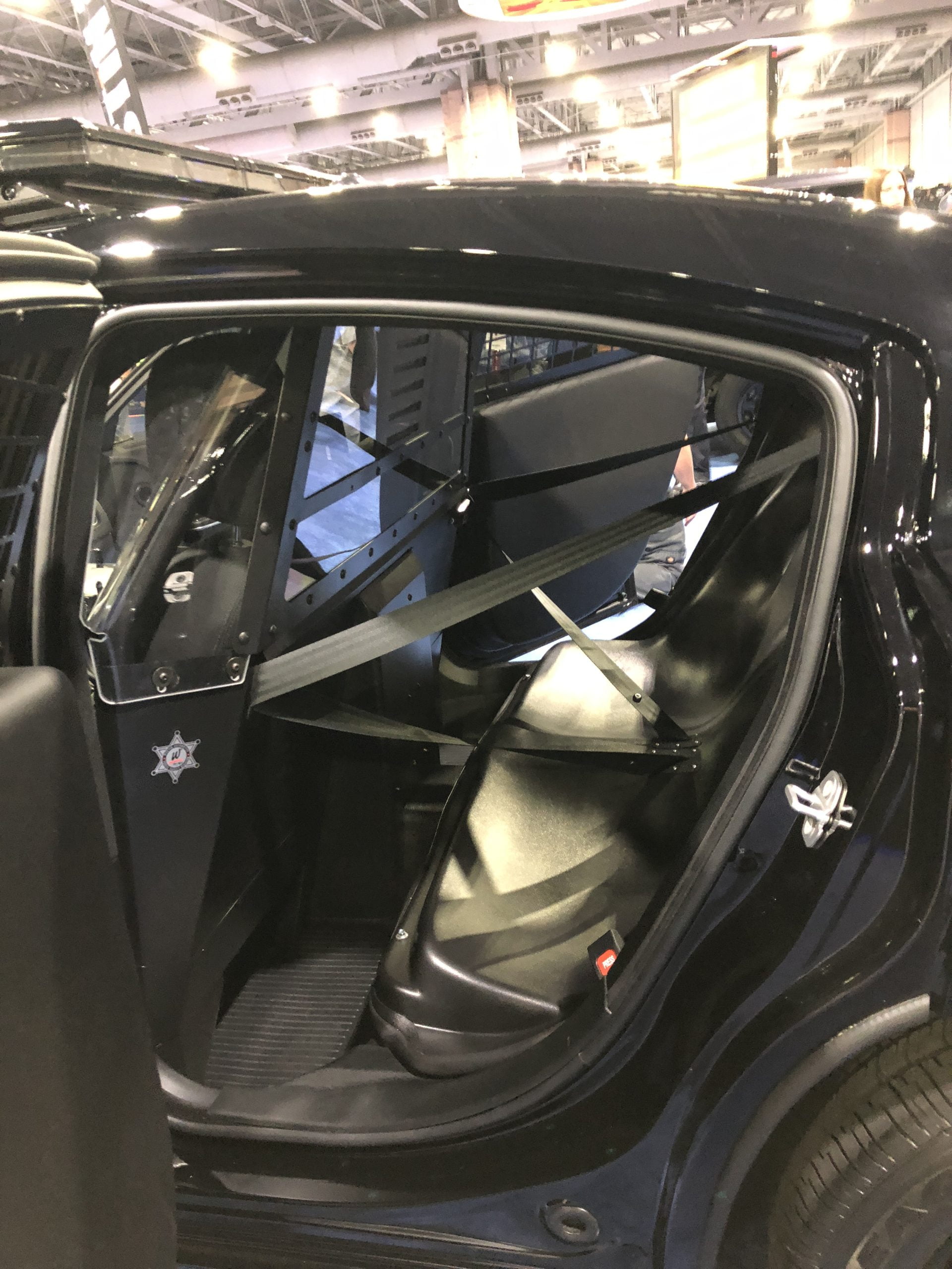 IMG_7398 seat belt systems Interior of a police vehicle, showing the rear seat with a partition divider and secured barriers on the windows and doors.